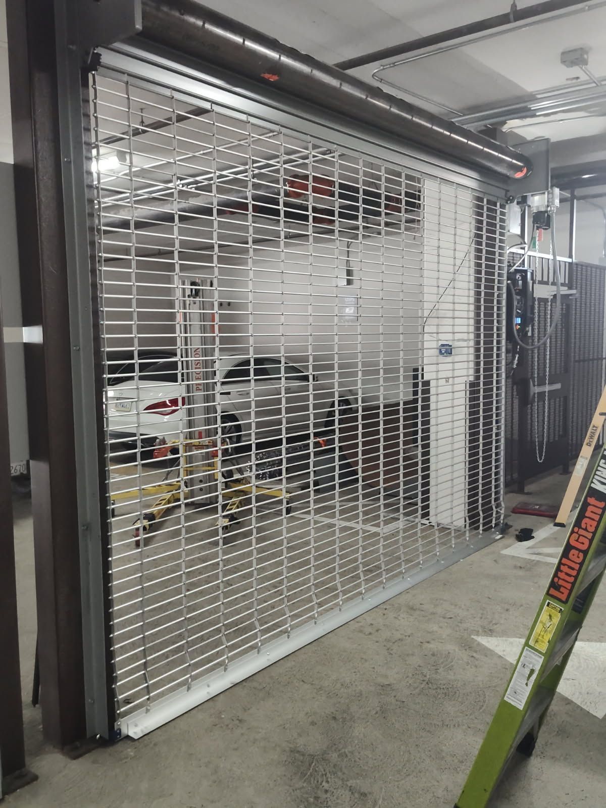 Metal security gate, partially open, revealing a parking garage with vehicles inside. A ladder rests nearby.