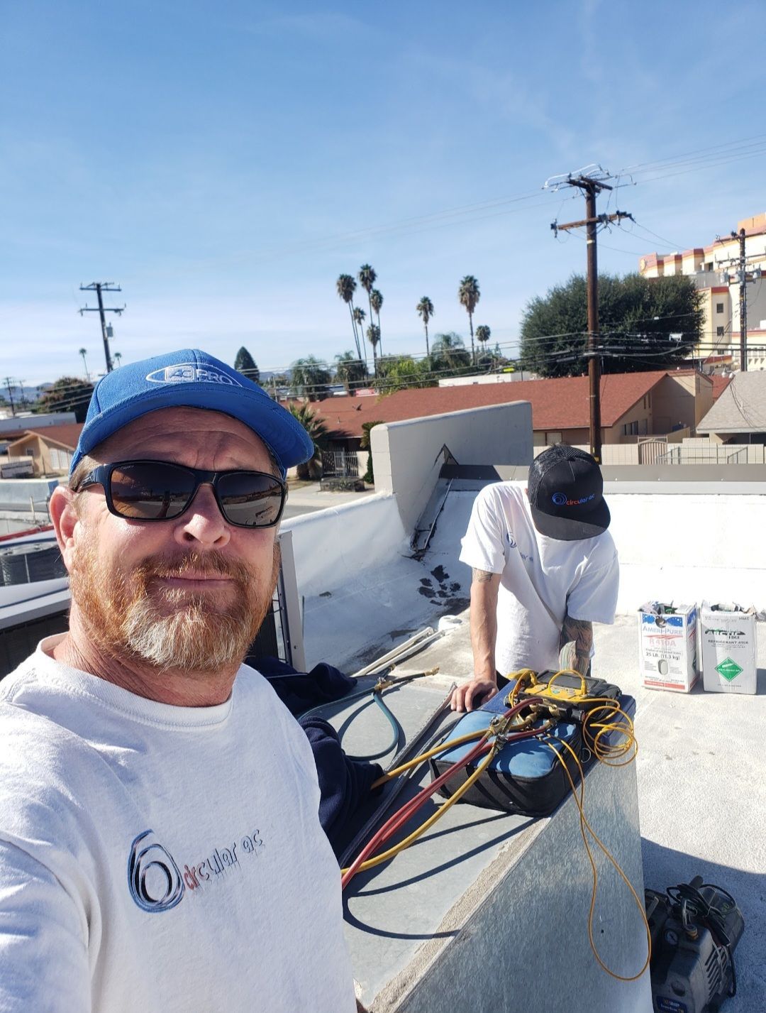 Two workers on a rooftop with cables and tools, one taking a selfie in bright sunlight.