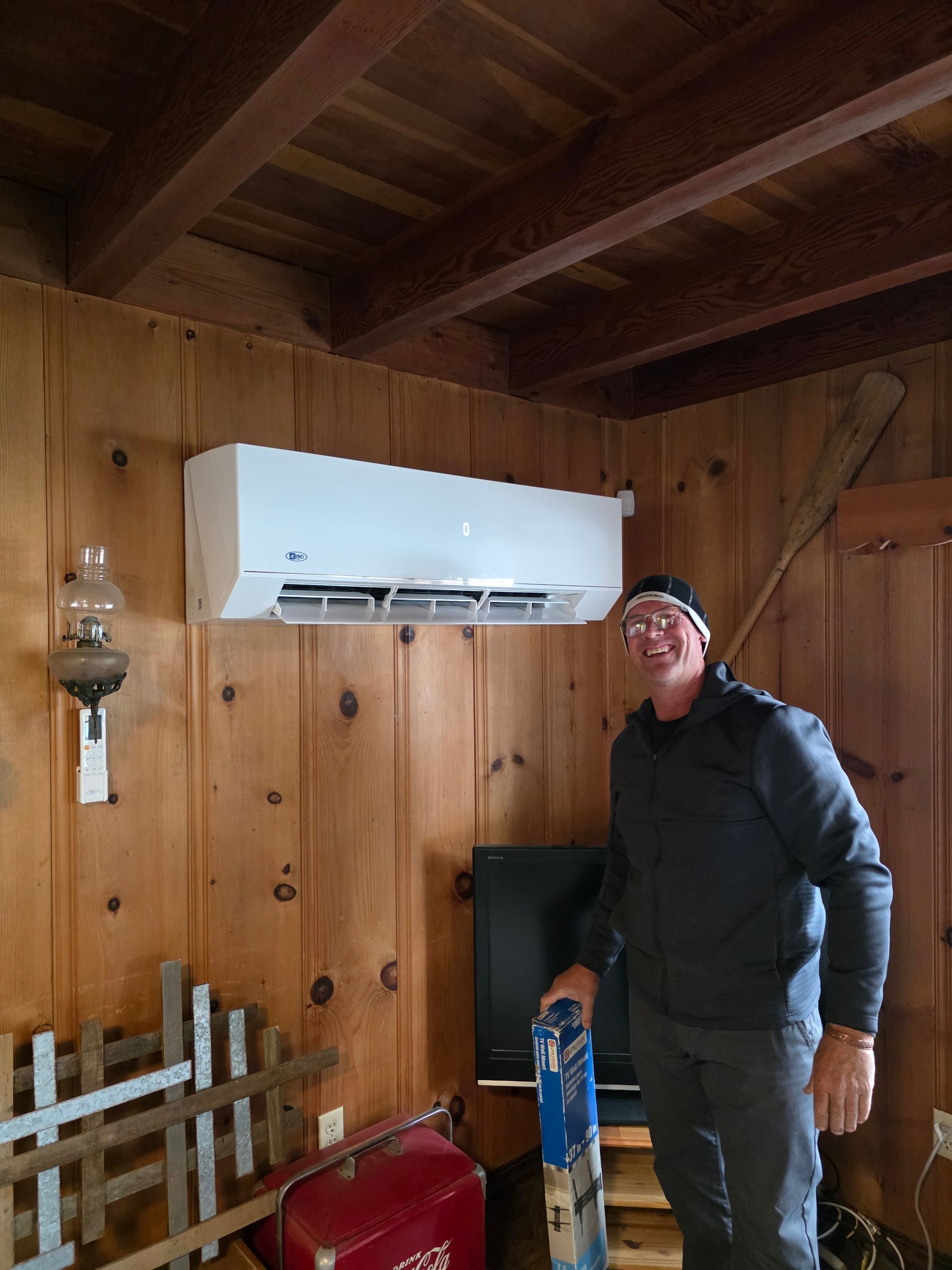 Man standing under a wall-mounted air conditioner in a wood-paneled room, holding a blue tool.