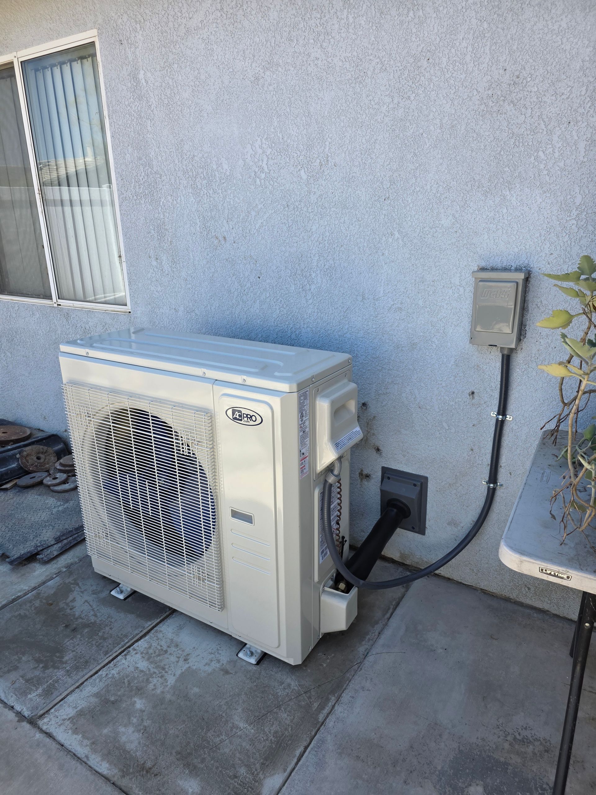 Outdoor air conditioning unit beside a stucco wall and utility box on a concrete patio.