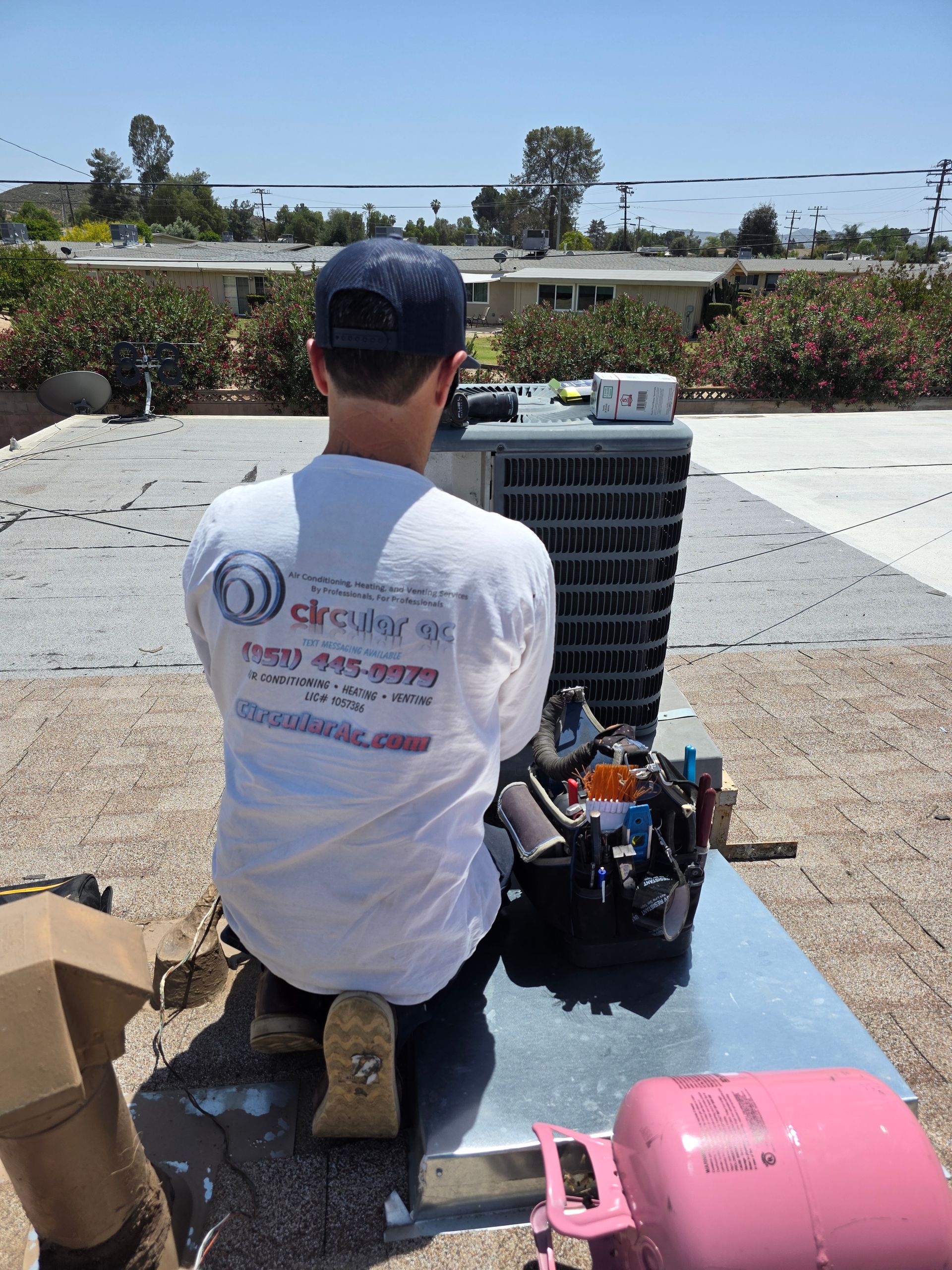 Technician kneels beside outdoor HVAC unit, working on equipment in a sunny yard.