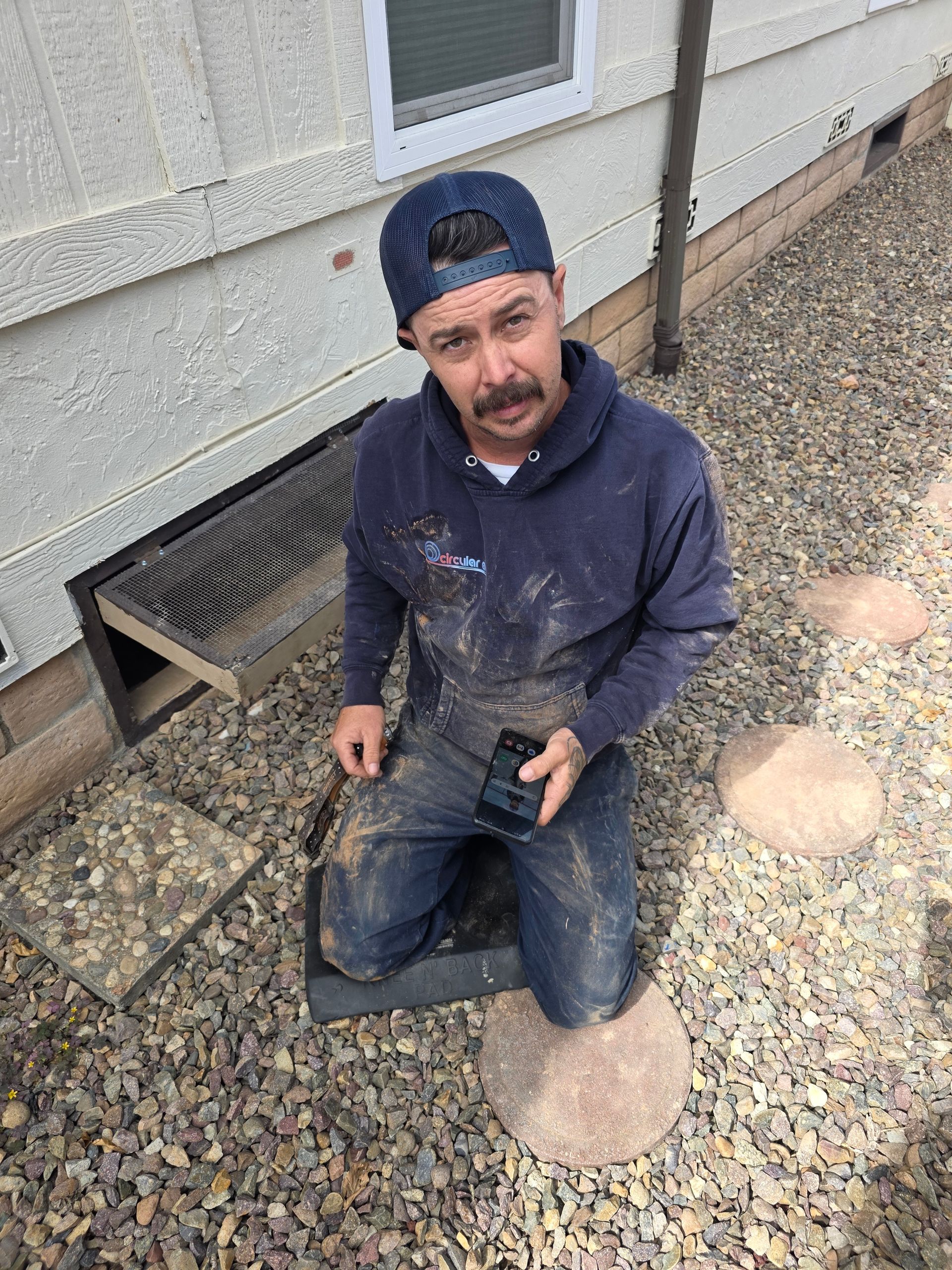 Worker kneeling by a house foundation drain, holding tools in a gravel area with a serious expression