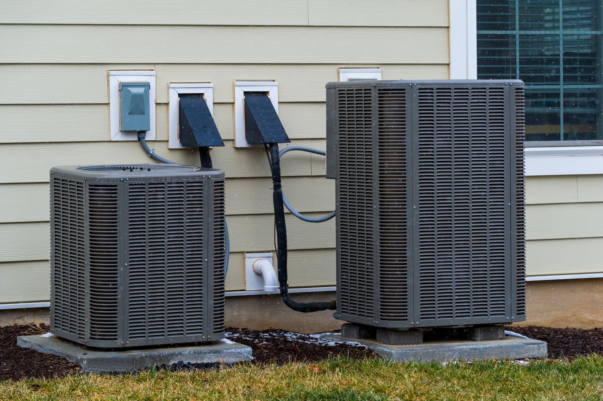 Two outdoor HVAC units beside a house wall, with electrical boxes and a window nearby.