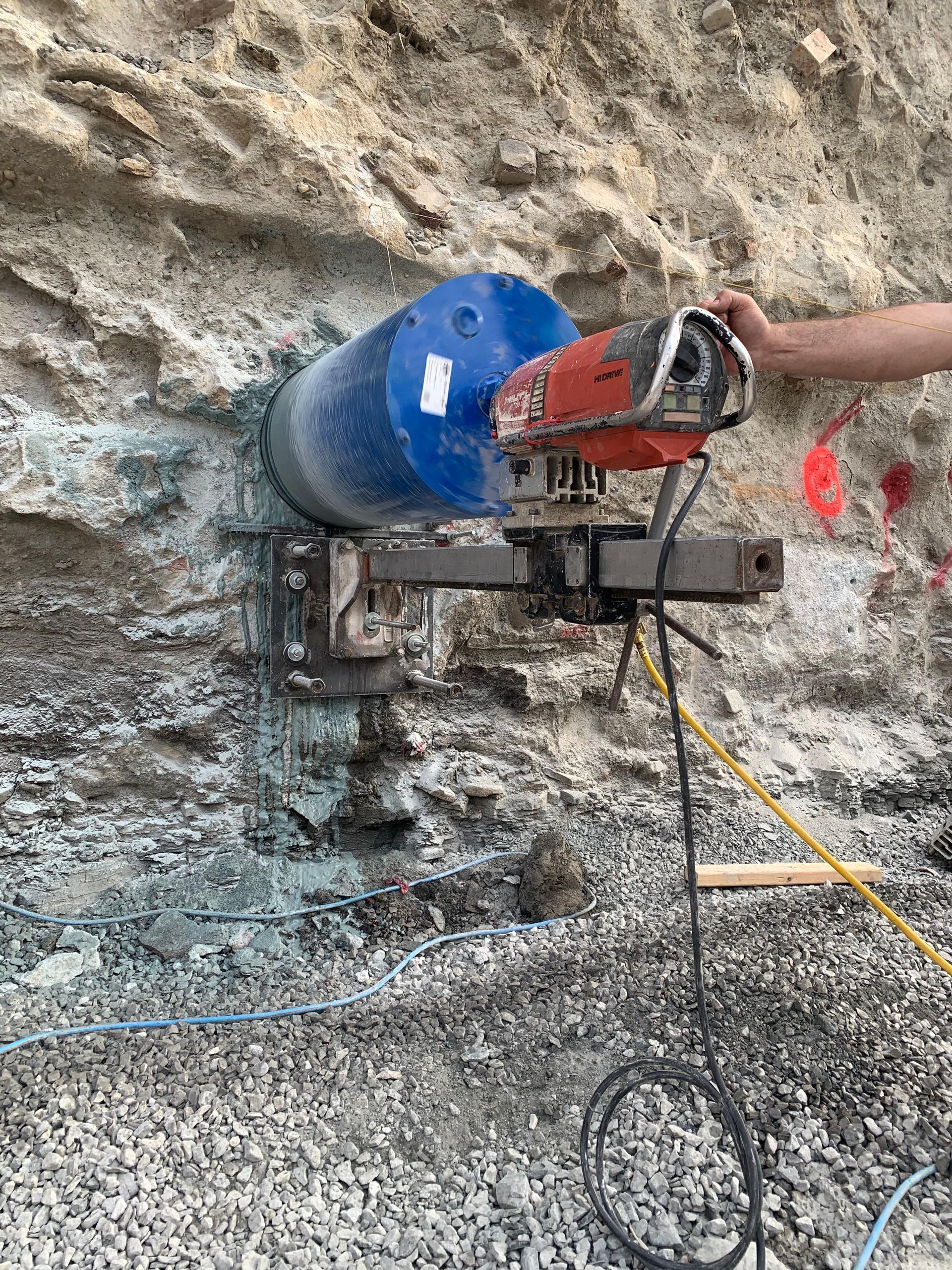 Core drilling machine cutting into a gray stone wall, blue and red components, outdoor setting.