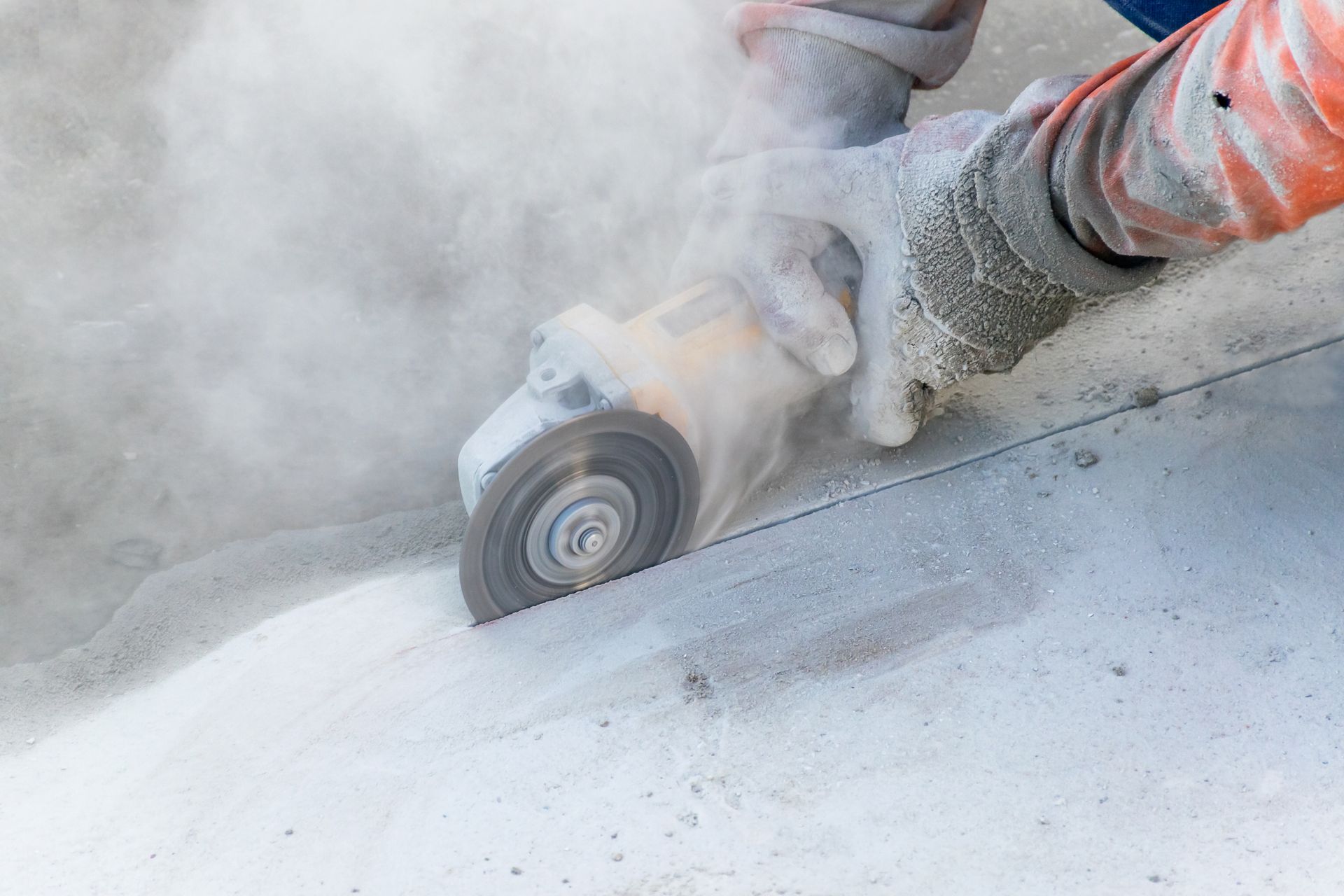 Person wearing gloves using a power saw, creating a large dust cloud while cutting concrete.