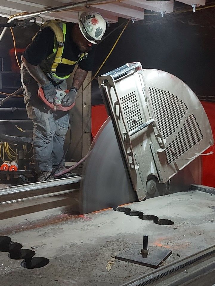 A worker in safety gear uses a large circular saw to cut into a concrete surface in an industrial setting.