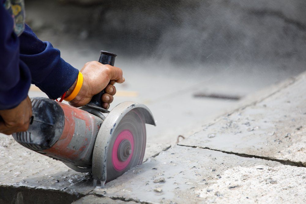 A person wearing a blue sleeve uses a handheld power saw with a pink-centered blade to cut through a concrete surface.