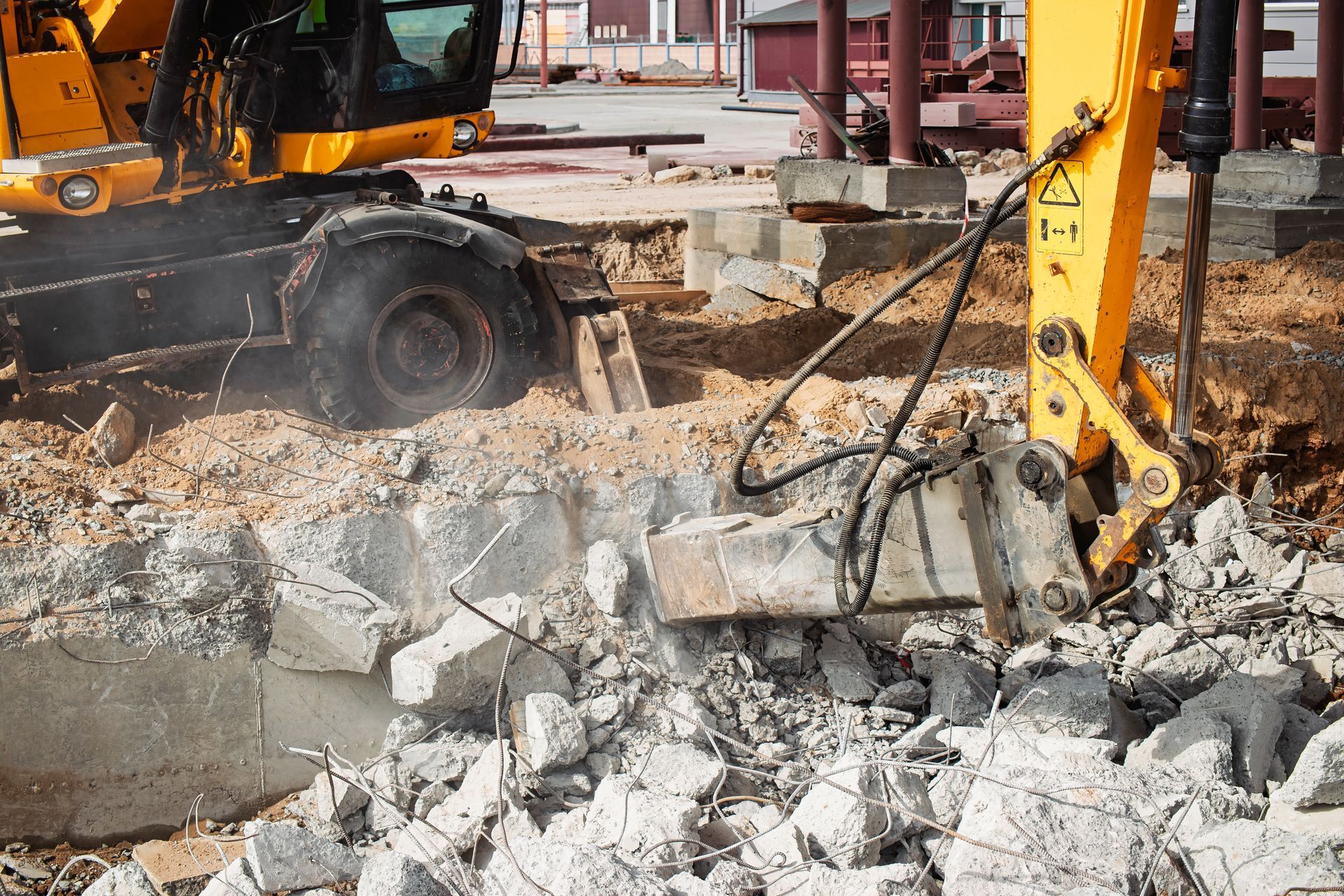 A yellow excavator breaks up concrete on a construction site, crushing debris with its metal attachment.