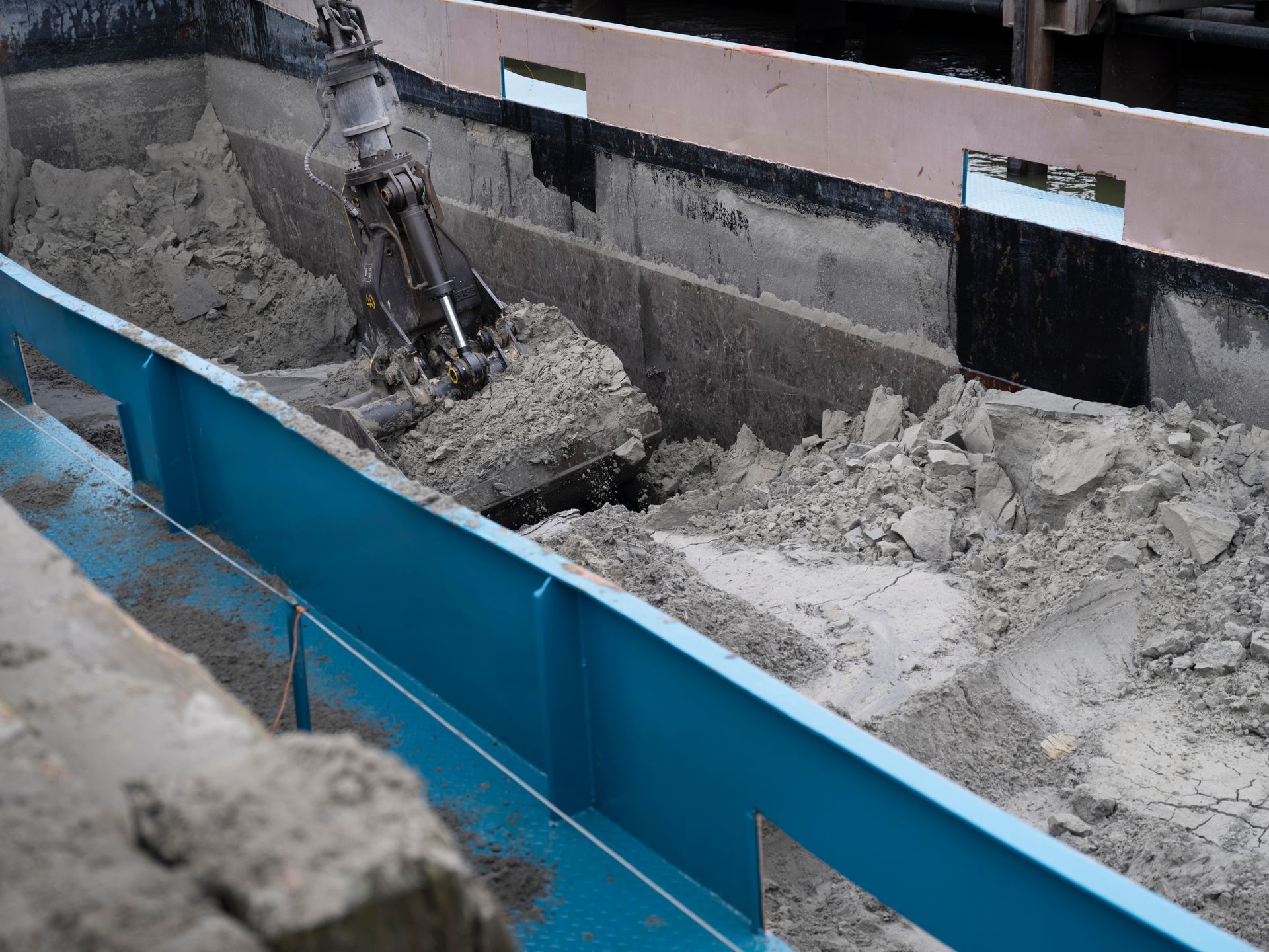 Excavator arm removing debris from a blue-edged trench, showing concrete and soil in a construction setting.