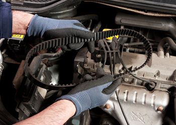 A man wearing gloves is holding a timing belt over a car engine.