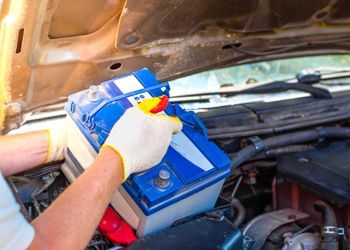 A person is holding a car battery under the hood of a car.