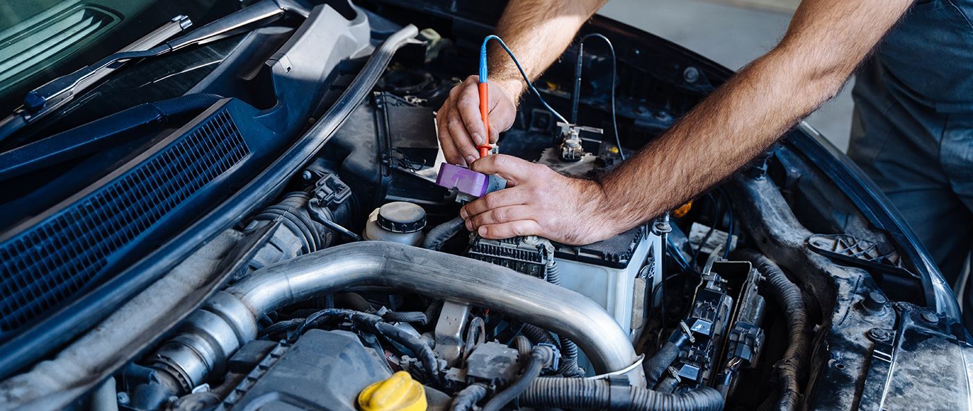 A mechanic uses a multimeter probe to test a car battery under the open hood of a vehicle.