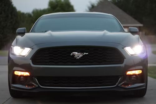 Dark gray Ford Mustang with bright headlights, parked in front of a house.