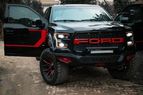 Black Ford Raptor truck with red accents, large tires, and open door. Parked on a dirt path.