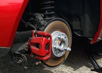 Red car's rear brake disc and caliper exposed. Rusty rotor, red caliper, wheel hub, jack underneath