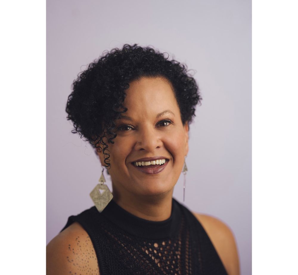 Woman with dark curly hair and dangling earrings smiles; lavender background.
