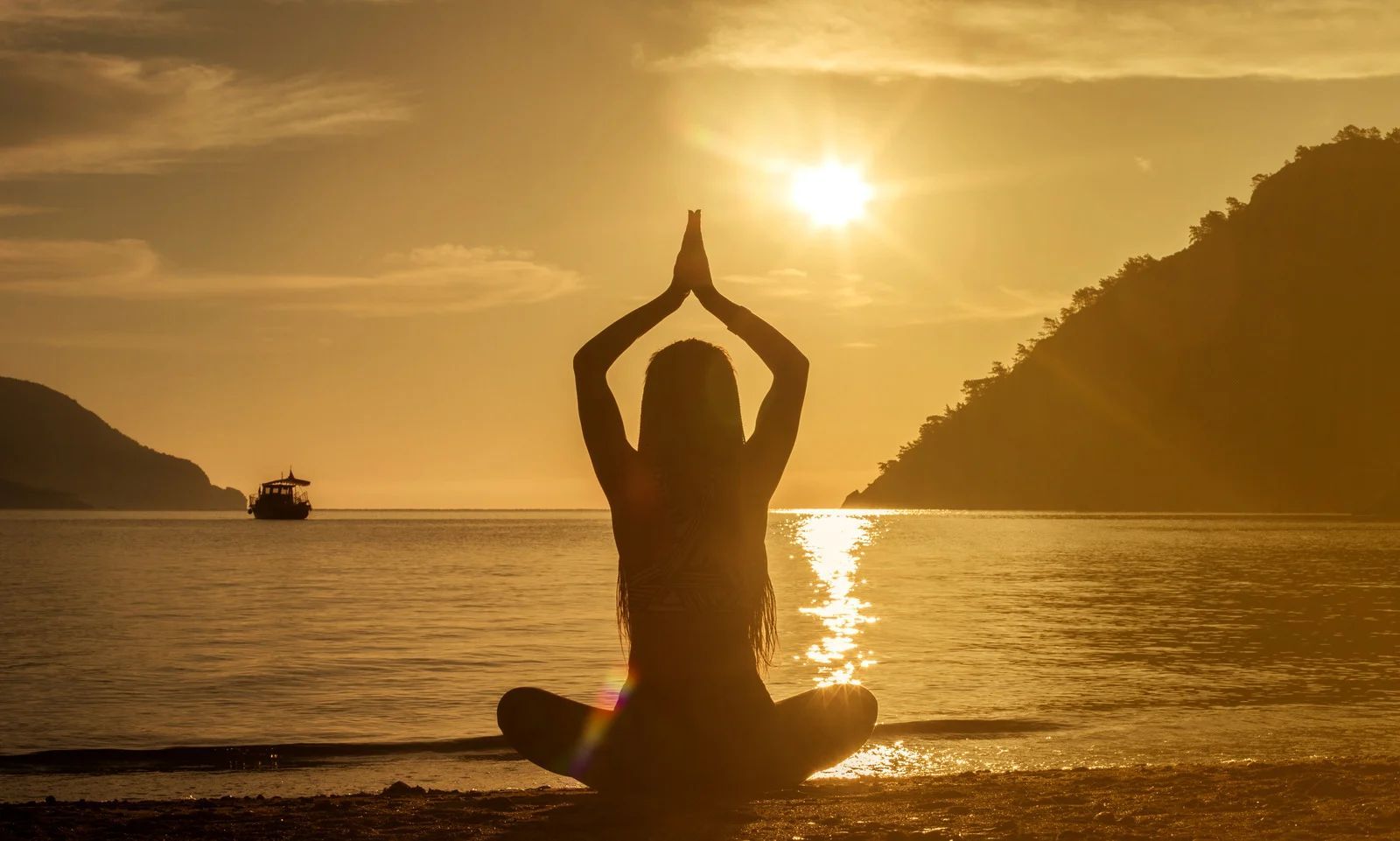 Silhouette of a person meditating on a beach at sunset with hands raised, ocean and boats in the background
