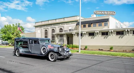 Vintage hearse on road in front of a building with "SHAPIRA & SONS" sign.