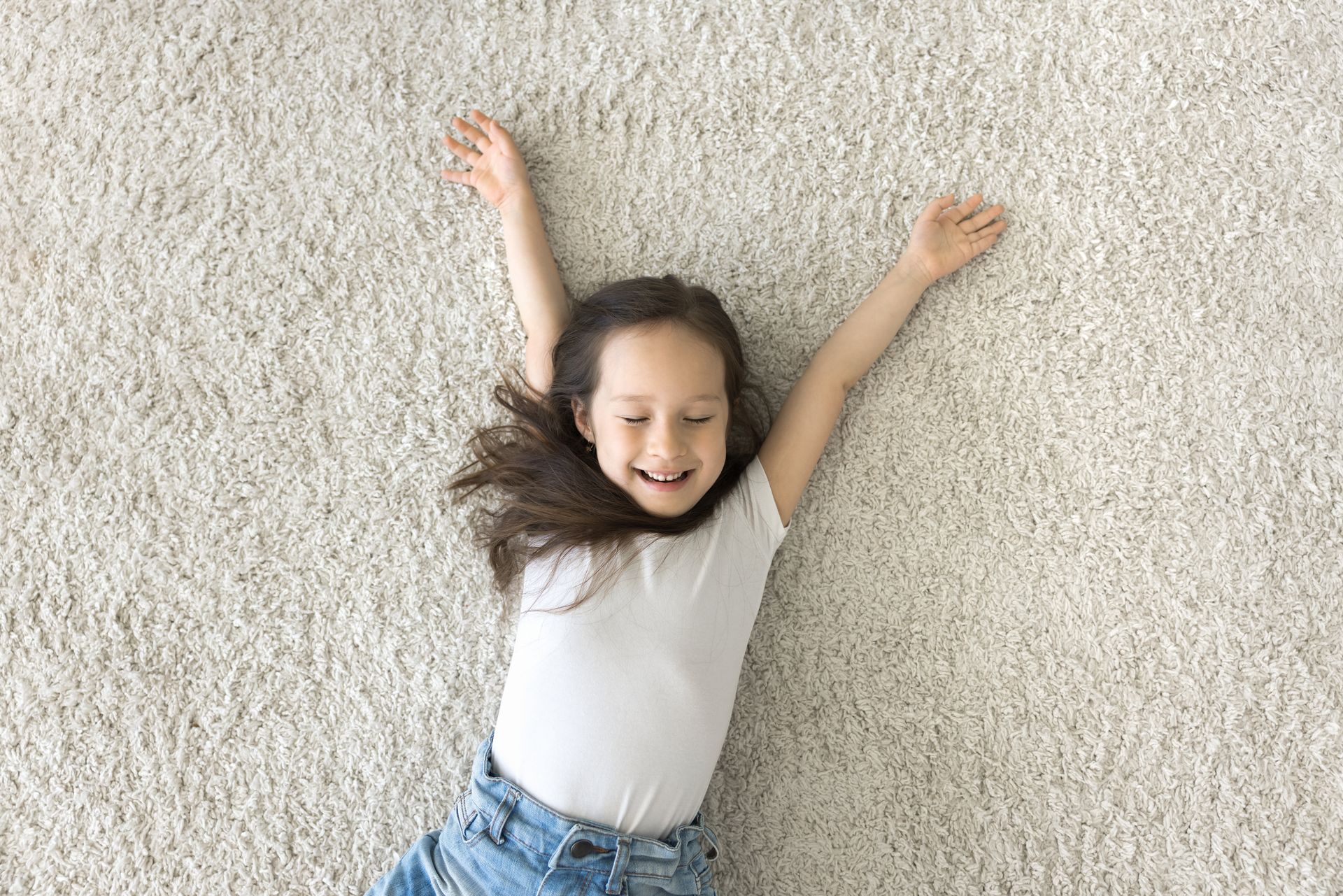 Smiling child lying on a light carpet with arms raised, wearing a white shirt and blue jeans