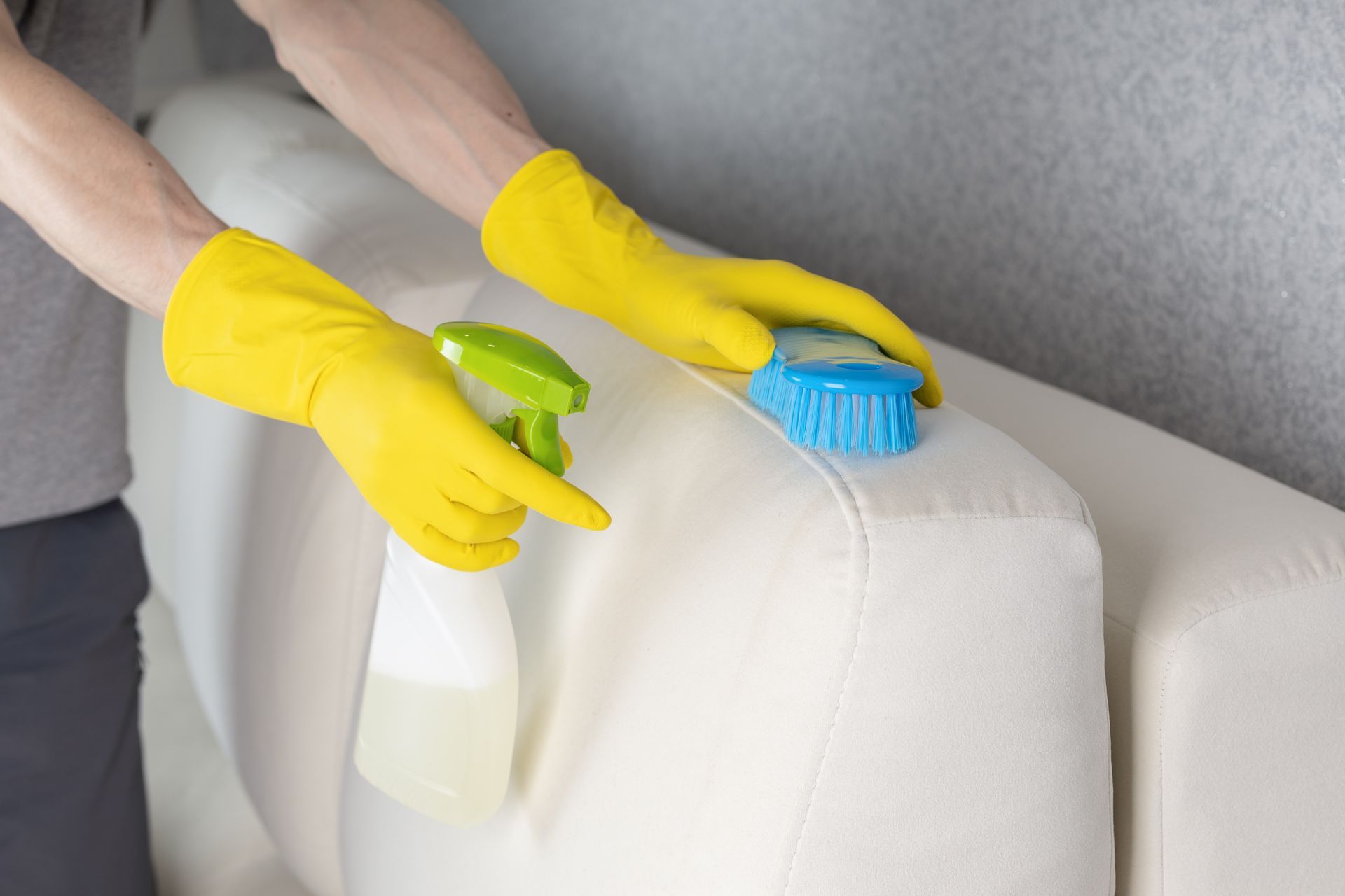 Person in yellow gloves cleaning a white couch with spray bottle and brush