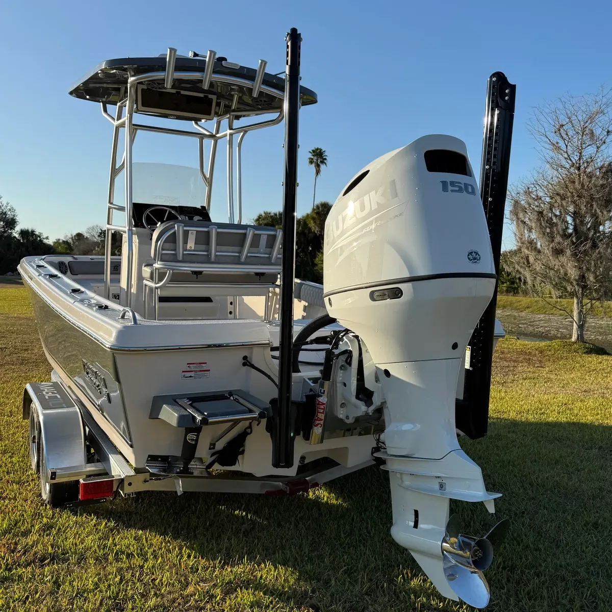 White center-console fishing boat on a trailer with large outboard motor, parked on grass by the water