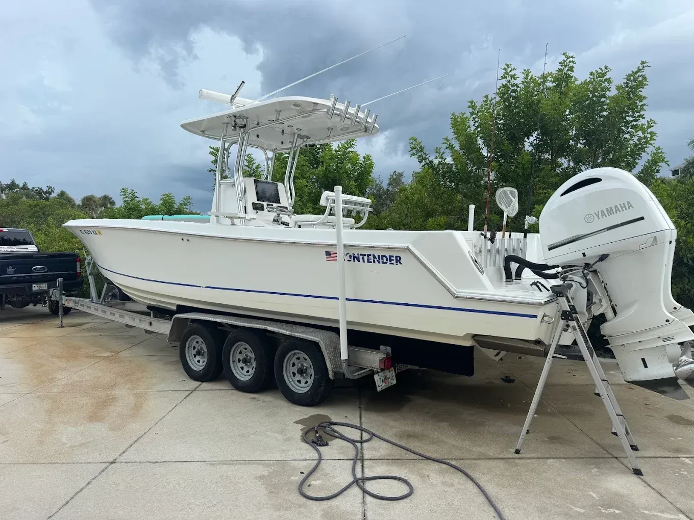 White center-console boat on a trailer with twin outboards, parked on wet pavement under cloudy skies.