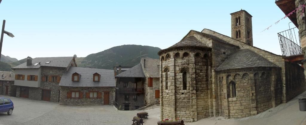 Una pintoresca plaça d'un poble europeu amb edificis de pedra i una església al fons, un cotxe blau i un arbre.