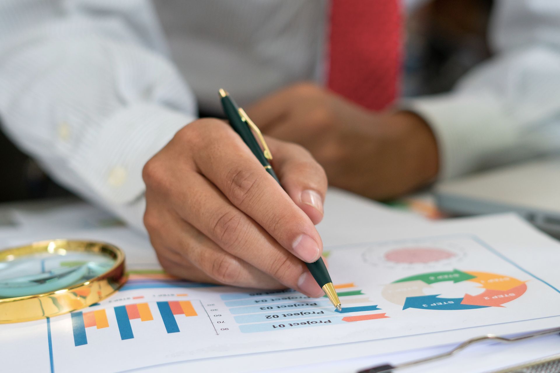 Hand holding a pencil over printed charts and graphs on a desk, with a magnifying glass nearby