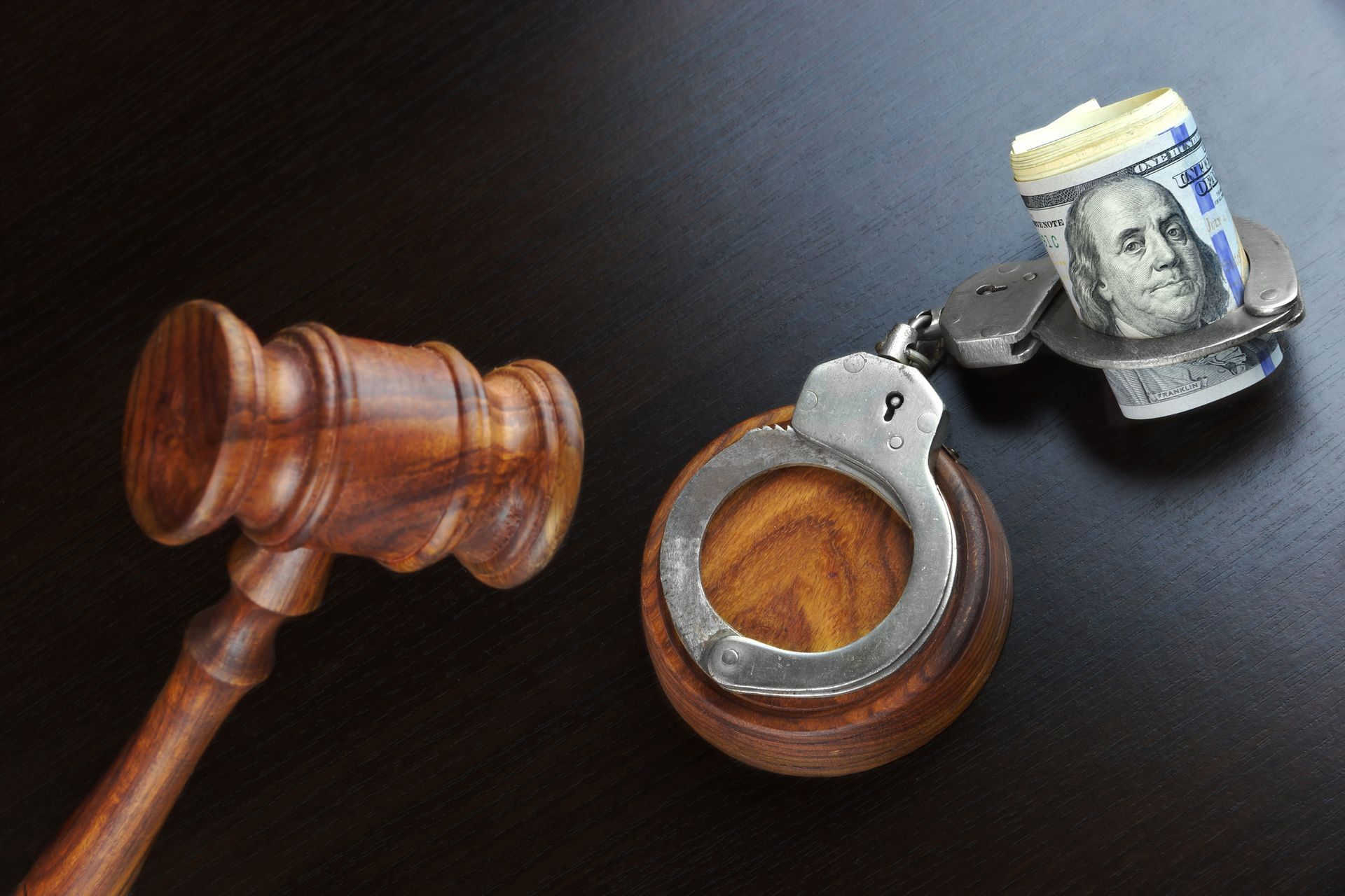 Judge’s gavel, handcuffs, and a rolled dollar bill on a dark surface.