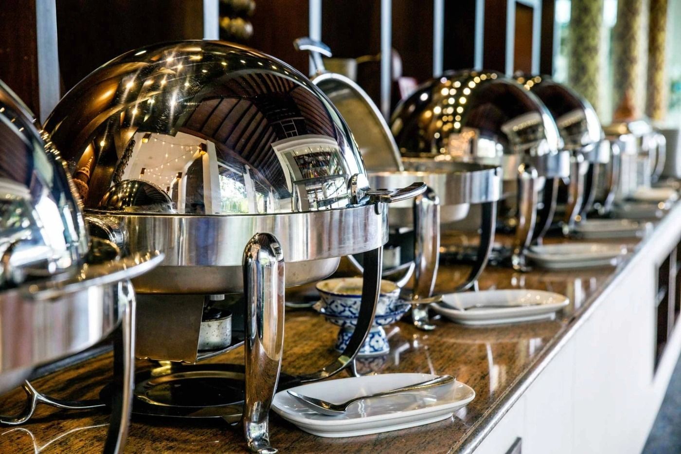 Buffet table with polished silver chafing dishes and serving utensils lined up in a dining room