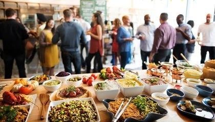 Buffet table with assorted dishes in foreground and people chatting at a social gathering in the background