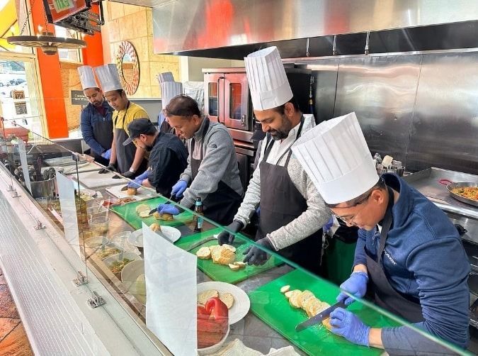 Chefs in tall white hats prepare dishes at a long counter in a bright commercial kitchen