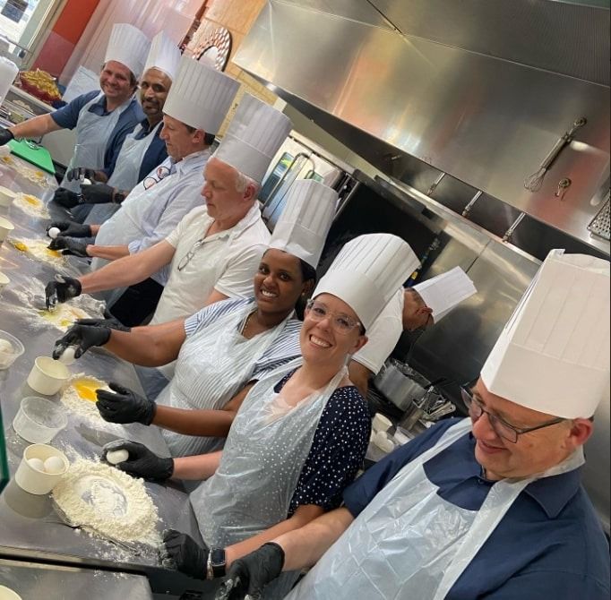 Chefs in white hats and aprons smiling while working together in a commercial kitchen with dough on the counter