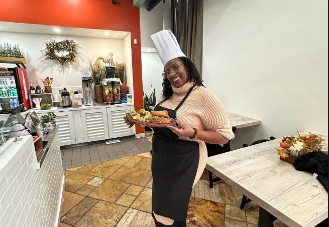 Woman in chef hat holding a tray in a bright kitchen, smiling beside a white table.