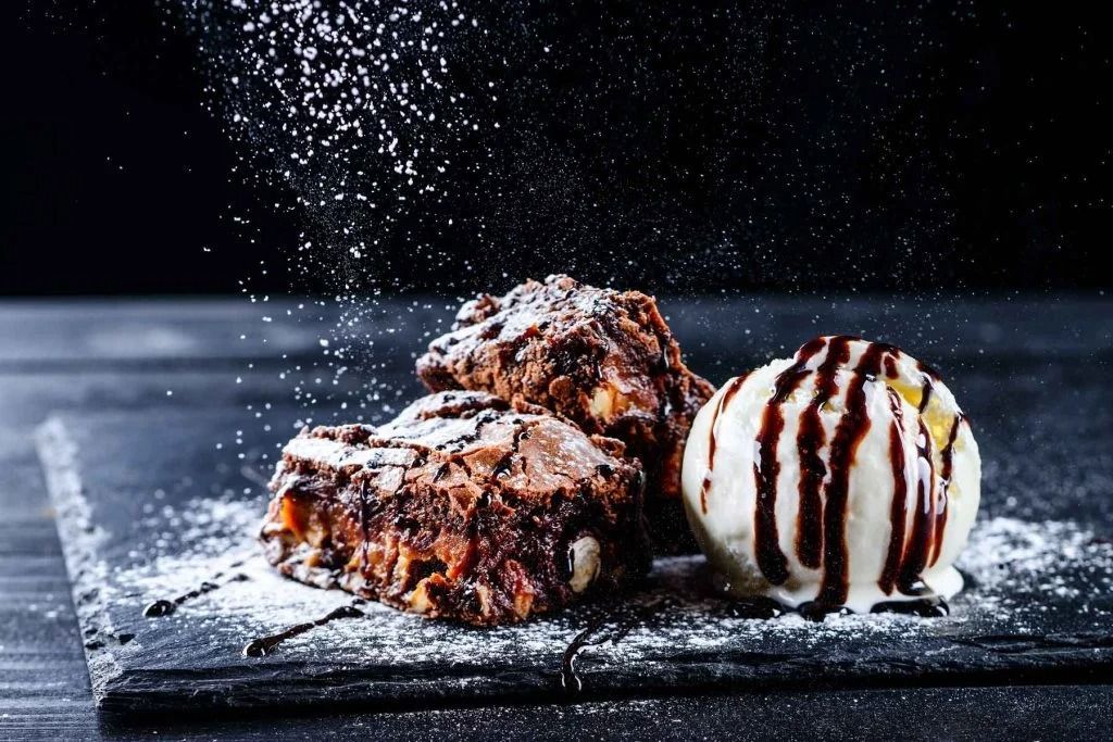 Chocolate brownie with vanilla ice cream, drizzled with sauce, on a dark table with powdered sugar falling around it