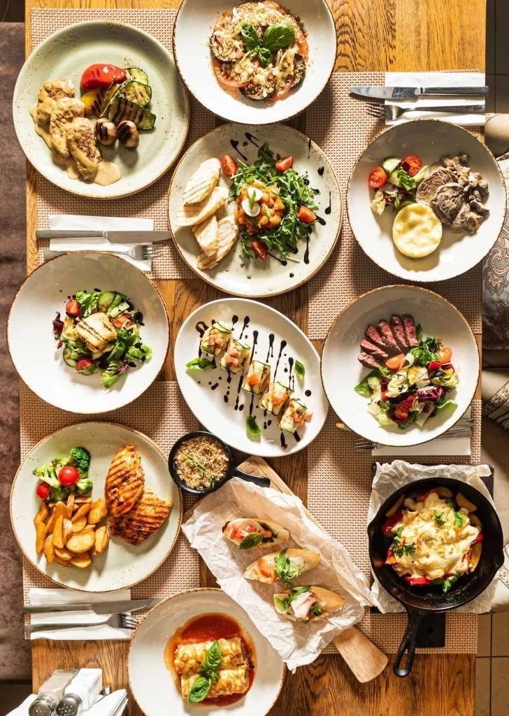 Overhead view of a table filled with assorted plated dishes, salads, pasta, and bread in warm light