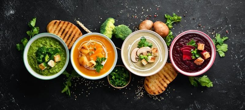 Four colorful bowls of soup on a dark table with bread, herbs, and mushrooms around them