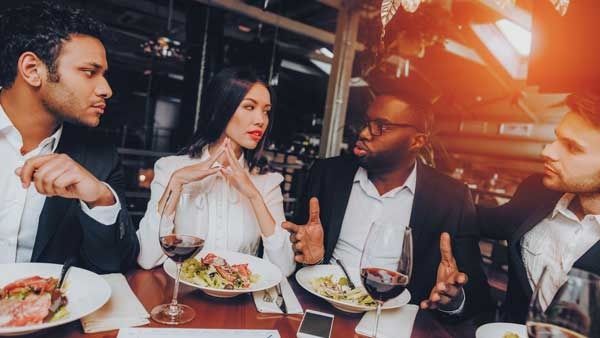 Four people dine and talk at a candlelit restaurant table with wine and plates of food.