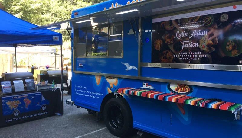 Blue food truck and tent at an outdoor market, with colorful food displays and a serving window.