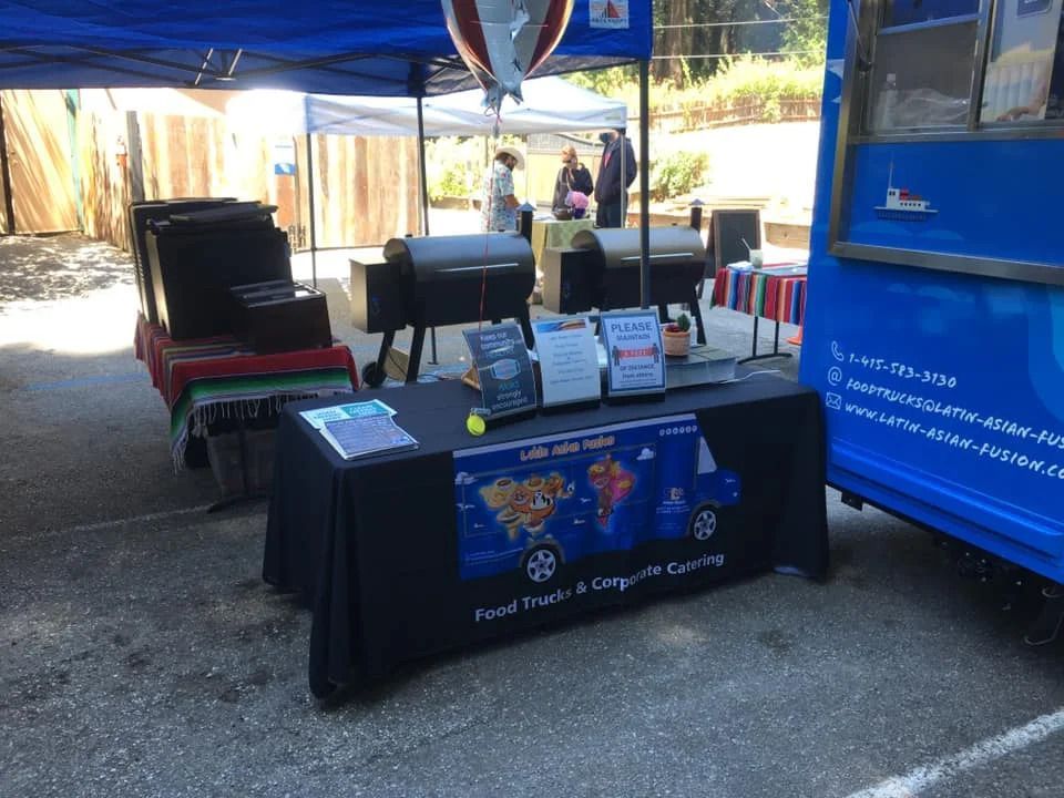 Booth under blue canopy with tables, signs, and a blue truck at an outdoor event area.