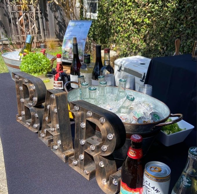 Outdoor party table with marquee letters, beer bottles, cups, and a tray of drinks in a sunny garden
