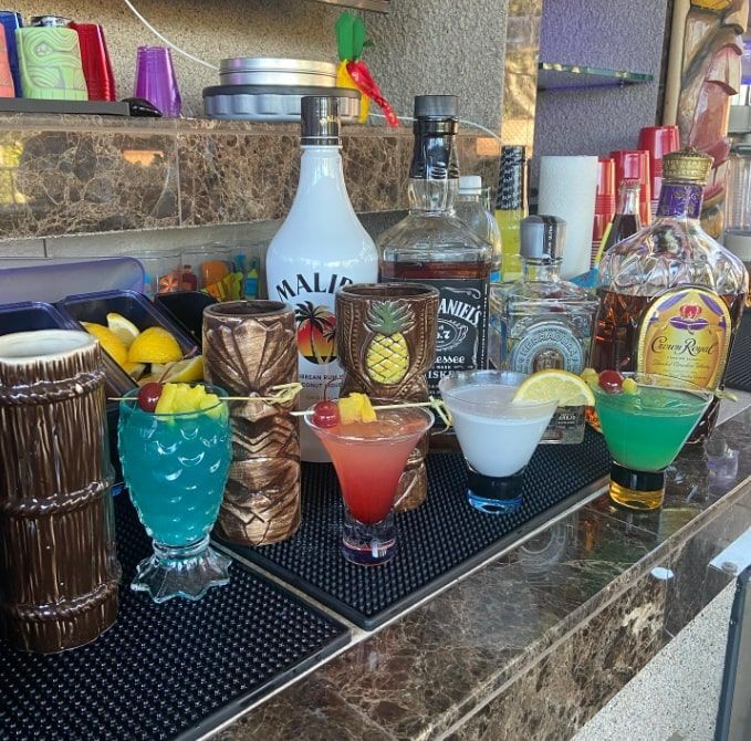Colorful cocktails lined on a bar counter, with bottles and tiki-style glassware in the background