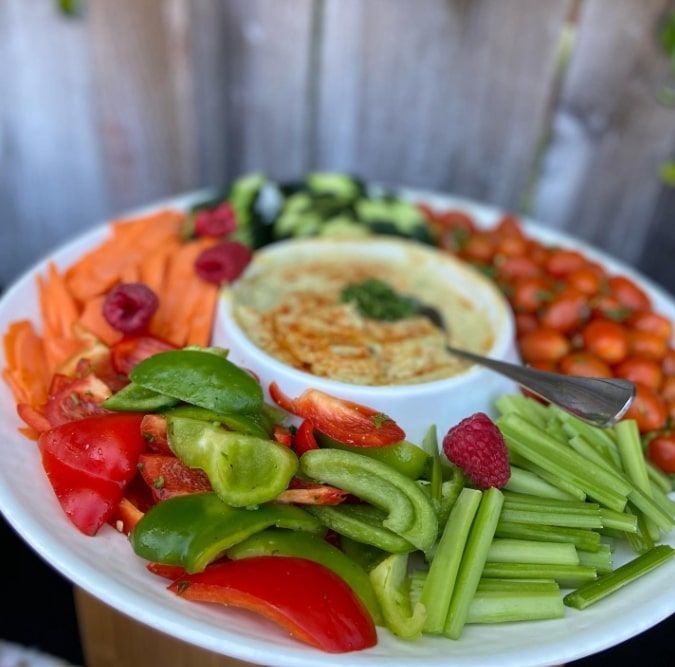 Fresh vegetables and hummus on a white platter, with red, green, and orange produce arranged around a bowl.
