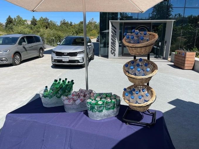 Outdoor vendor table with packaged snacks and green soda bottles under an umbrella, parked cars in background