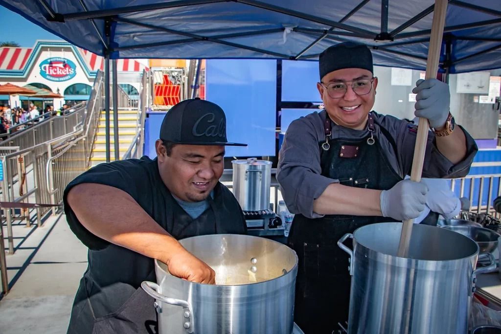Two cooks under a blue canopy stir large pots, smiling at an outdoor food stall.