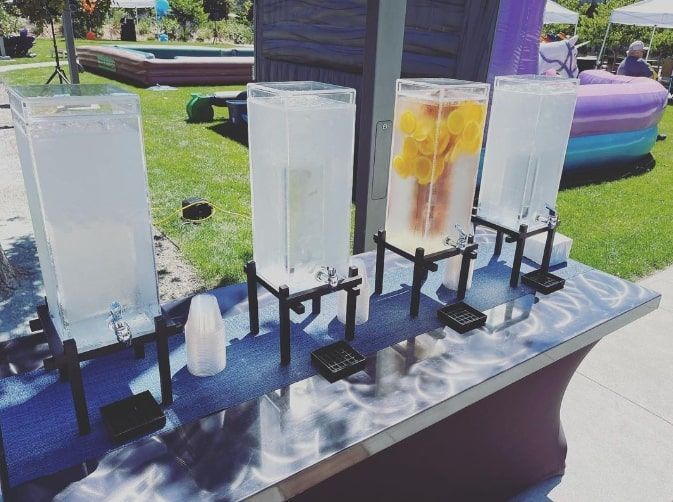 Four beverage dispensers on a metal table outdoors, two filled with iced drinks and fruit.