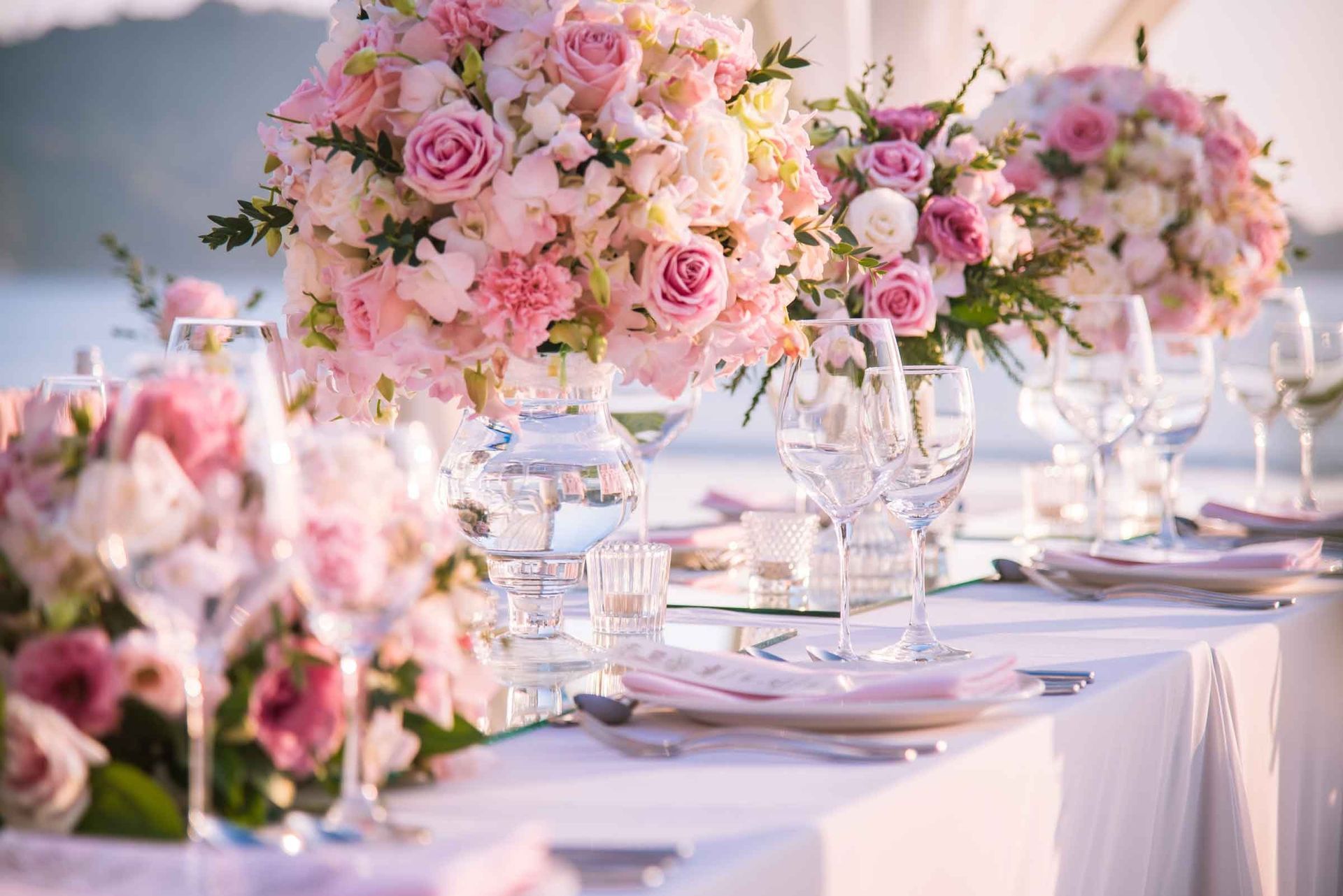Elegant outdoor table set with pink rose centerpieces, glassware, and white linens by the water