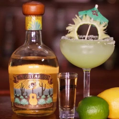 Tequila bottle, shot glass, and green cocktail garnished with citrus on a bar with limes and lemon.