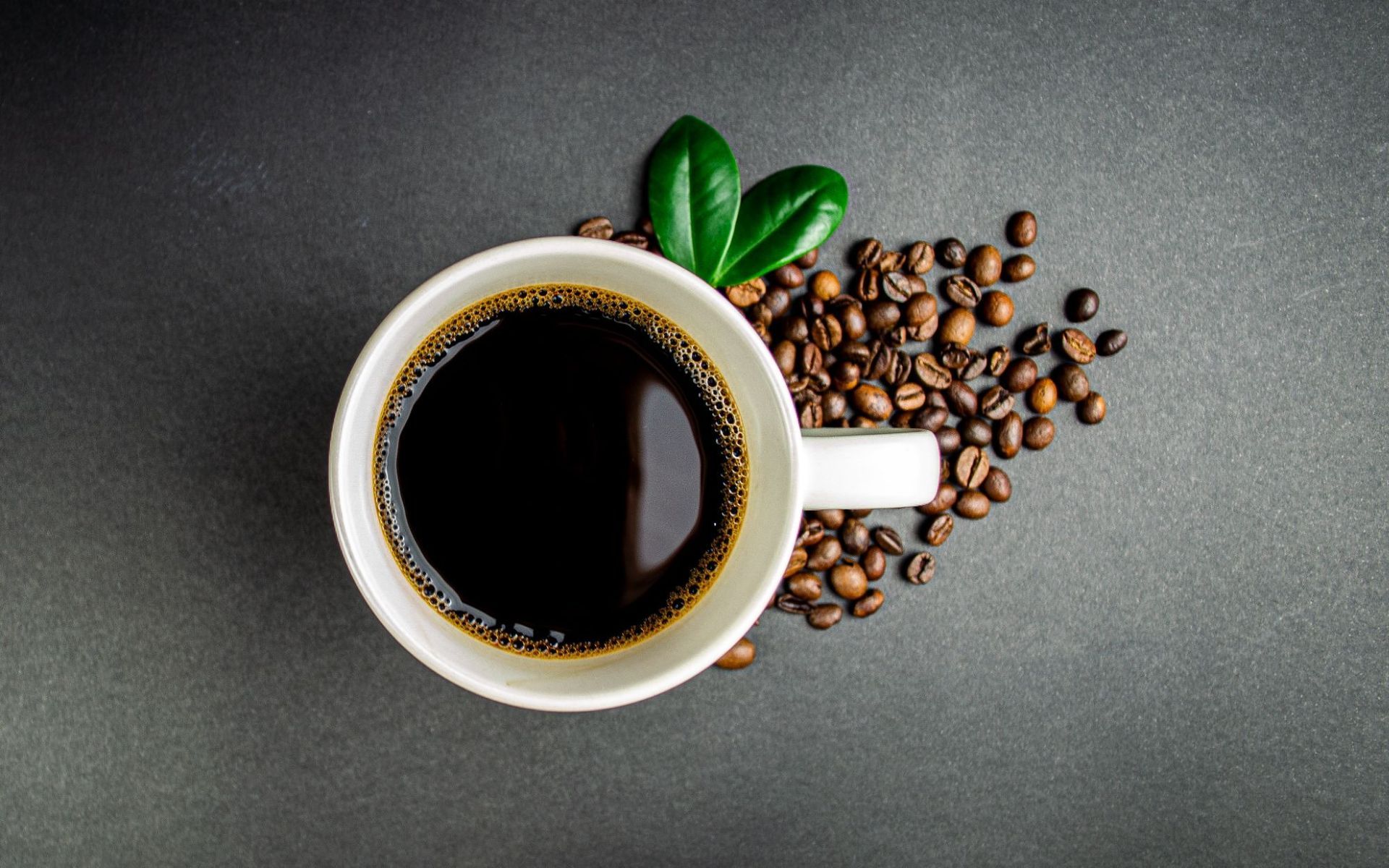 Top-down view of a black coffee cup with green leaves and scattered coffee beans on a gray surface