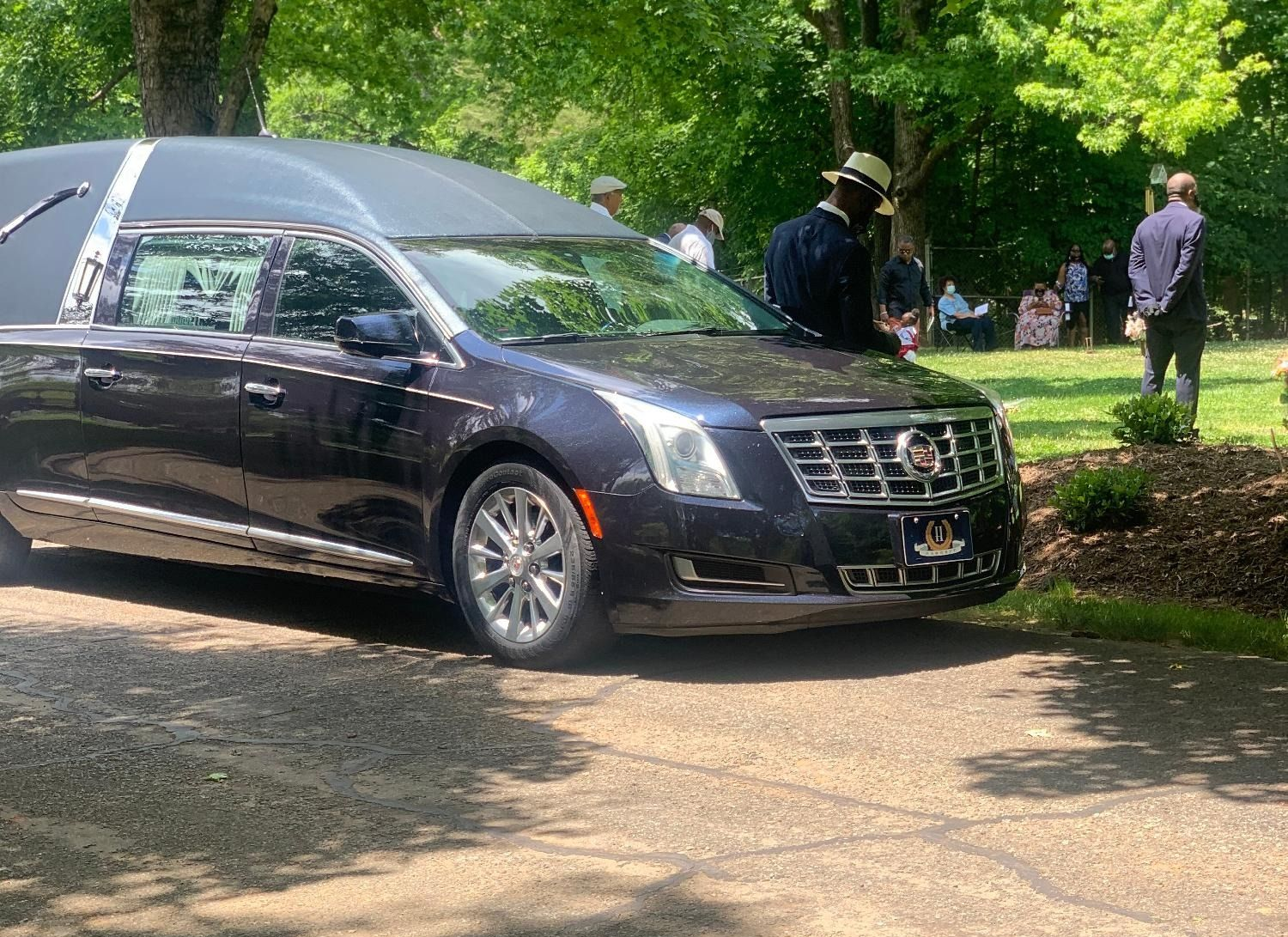 Hearse parked on a driveway, with people standing nearby in a park setting.